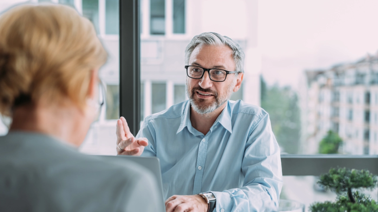 Ein Mann am Schreibtisch hinter einem Laptop im Gespräch mit einer gegenübersitzenden Frau.