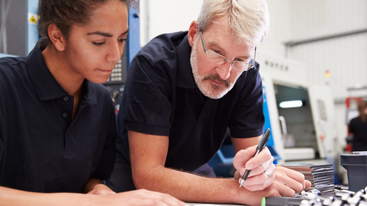 Junge Frau steht gemeinsam mit ihrem Ausbilder an einem Tisch und schaut in Unterlagen.