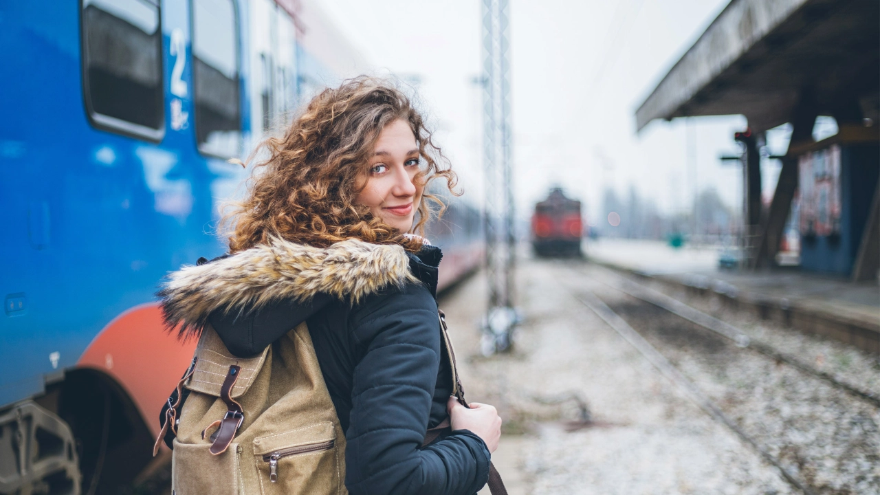 Junge Frau mit Rucksack auf dem Rücken steht vor einem Zug und blickt über ihre Schulter zurück in die Kamera.