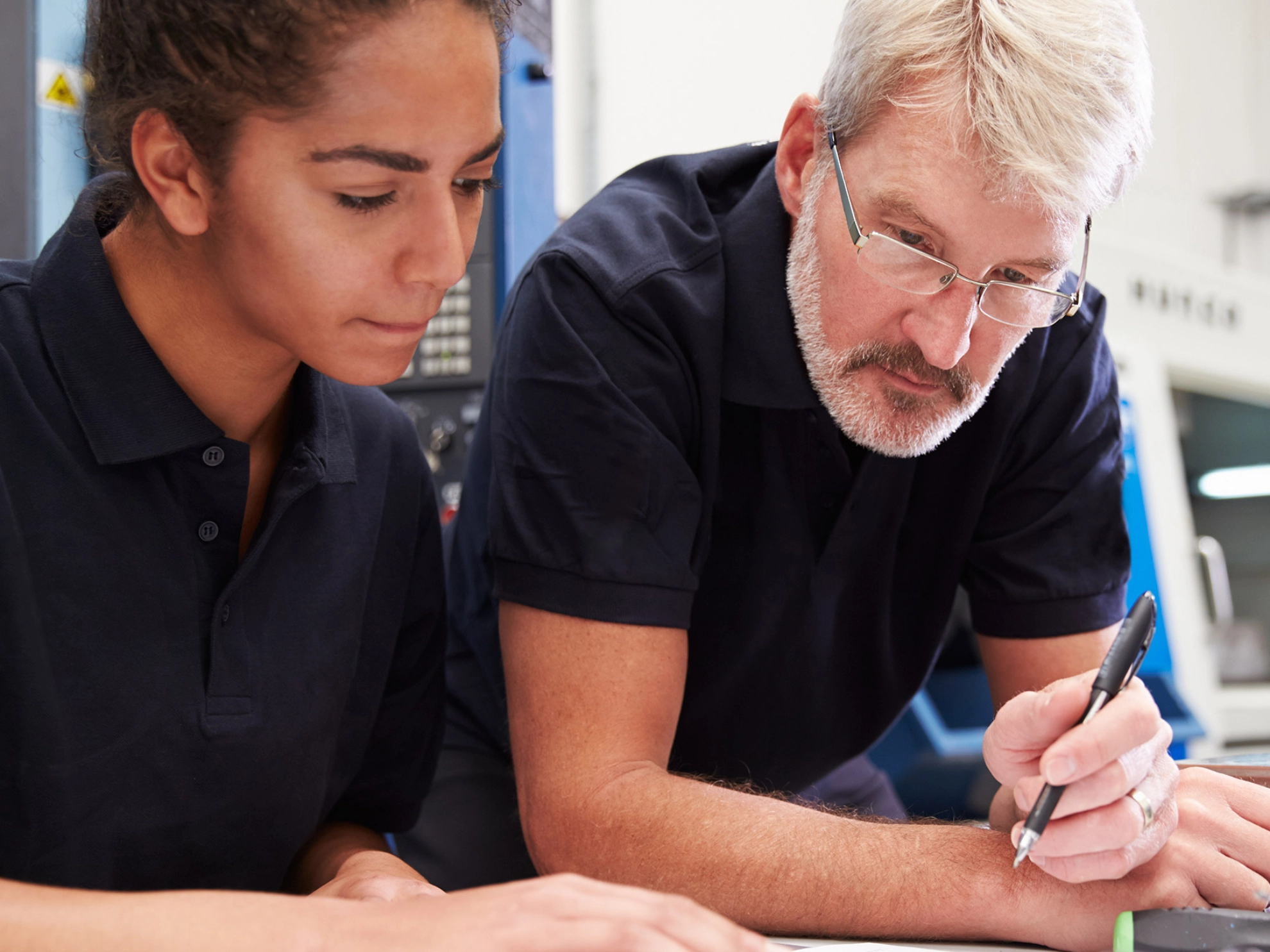 Junge Frau steht gemeinsam mit ihrem Ausbilder an einem Tisch und schaut in Unterlagen.