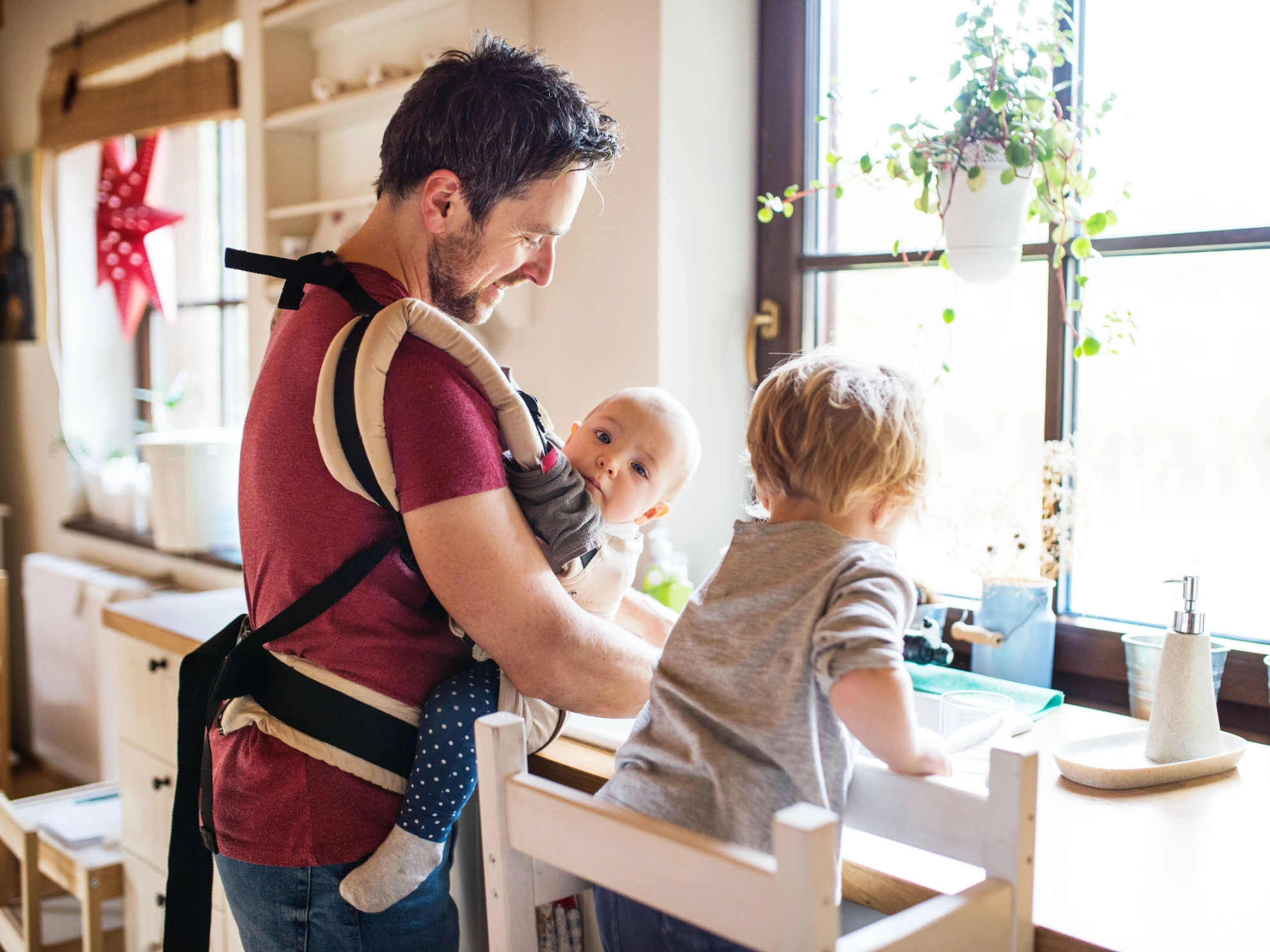 Vater mit Baby in Trage und zweitem Kind in der Küche