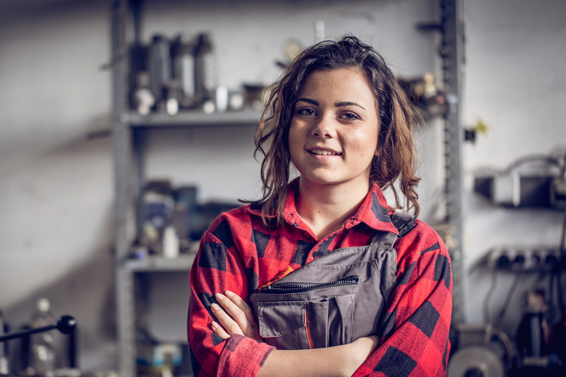 Junge Mechanikerin mit rotkariertem Hemd und Latzhose schaut mit verschränkten Armen selbstbewusst in die Kamera.