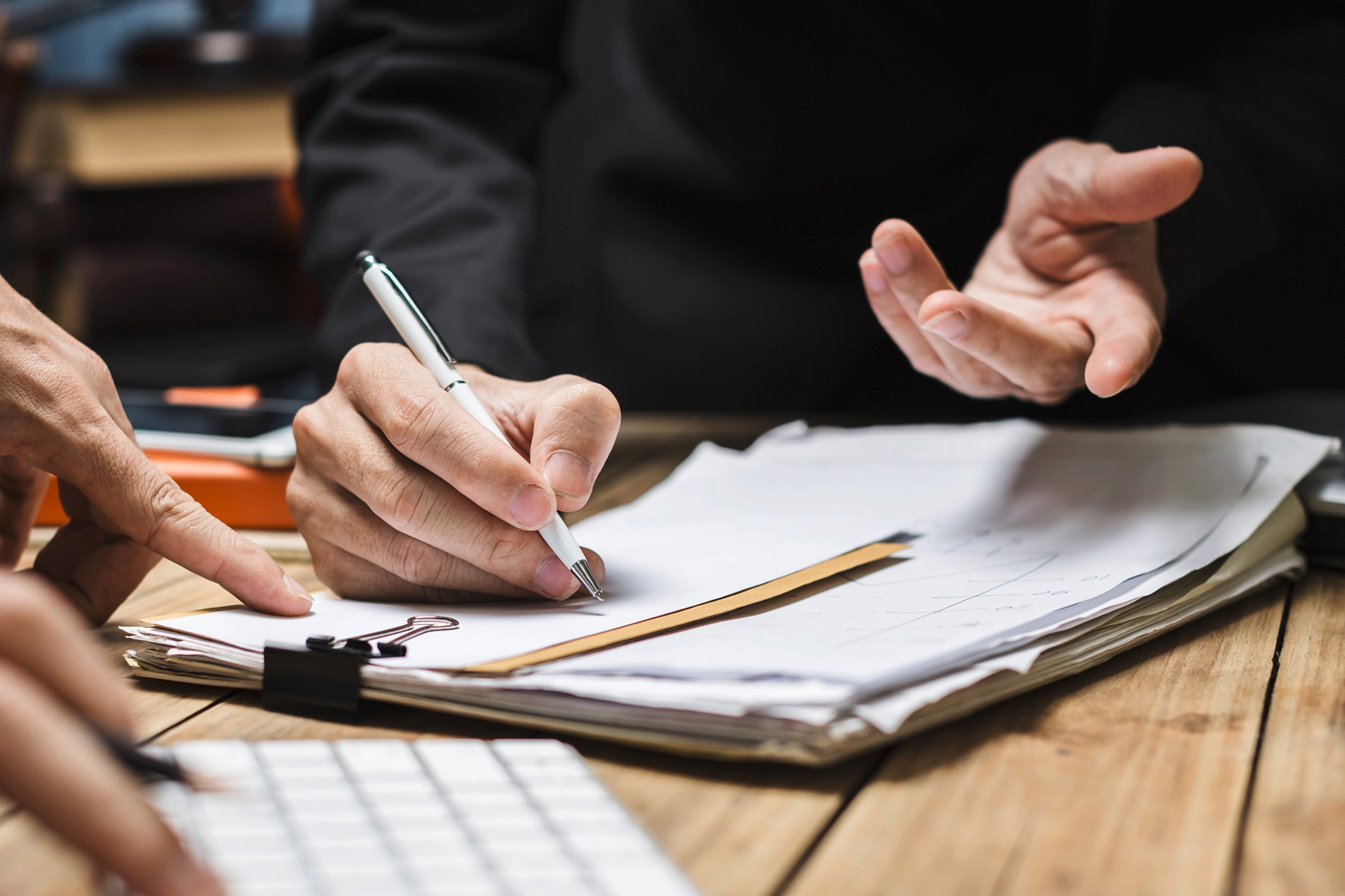 Ein macht Notizen auf Papier auf einem Klemmbrett. Eine andere Person tippt auf einer Tastatur.