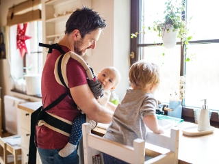 Vater mit Baby in Trage und zweitem Kind in der Küche