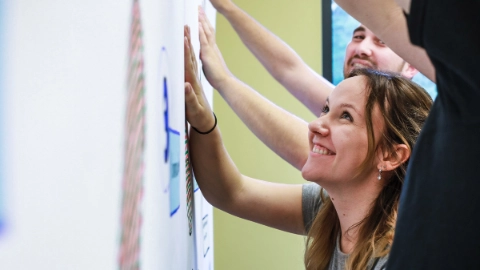 Junge Frau steht während eines Seminars mit weiteren Personen an einer Metaplanwand und schaut sich lächelnd die Aufschrift an.