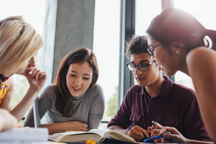 Vier Studierende blicken in Bücher, die vor ihnen auf dem Tisch liegen.